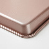 Close-up of a rose-coloured baking tray corner shows its finely textured surface and rolled rim against a white background.
