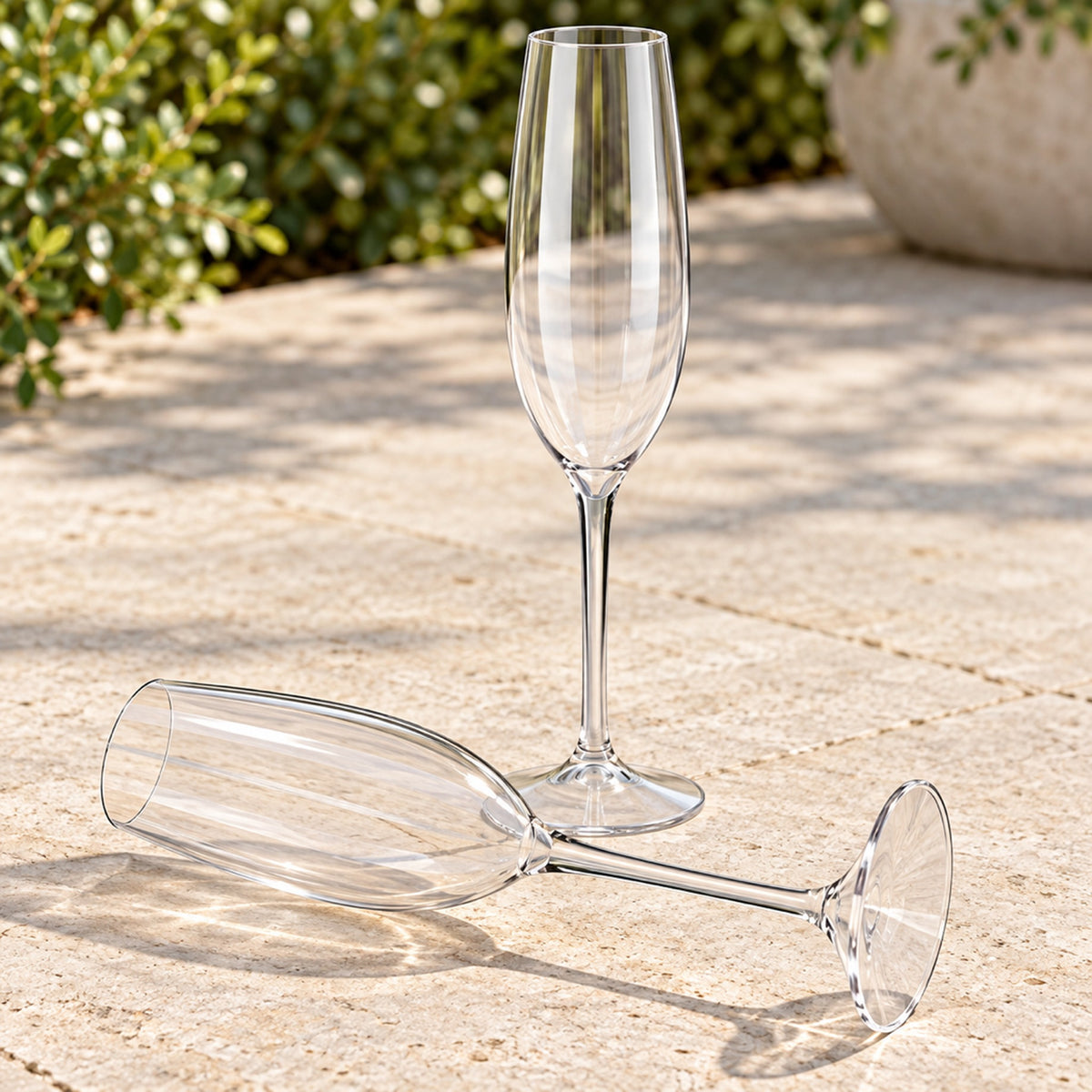 Clear Tritan plastic champagne flute lying intact on a stone patio beside an upright flute, showing it does not break