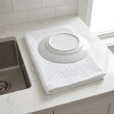 A white waffle-weave dish towel draped over a white plate, set on a white marble kitchen countertop next to a stainless steel sink.