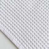 A close-up of a white waffle-weave fabric, likely a cotton kitchen towel or cloth, showing its textured grid pattern and folded corner.