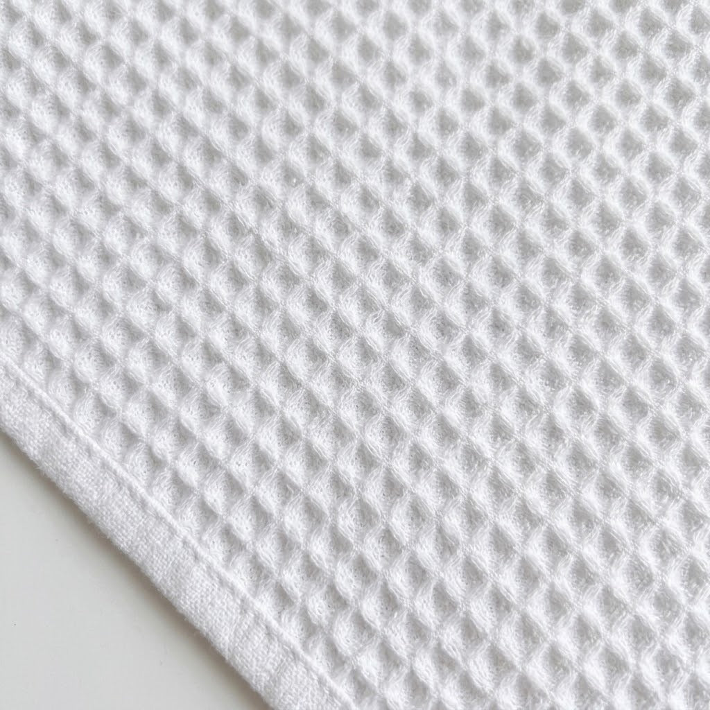A close-up of a white waffle-weave fabric, likely a cotton kitchen towel or cloth, showing its textured grid pattern and folded corner.