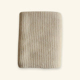 A folded beige textured blanket with a waffle or honeycomb knit pattern, shown against a solid off-white background.