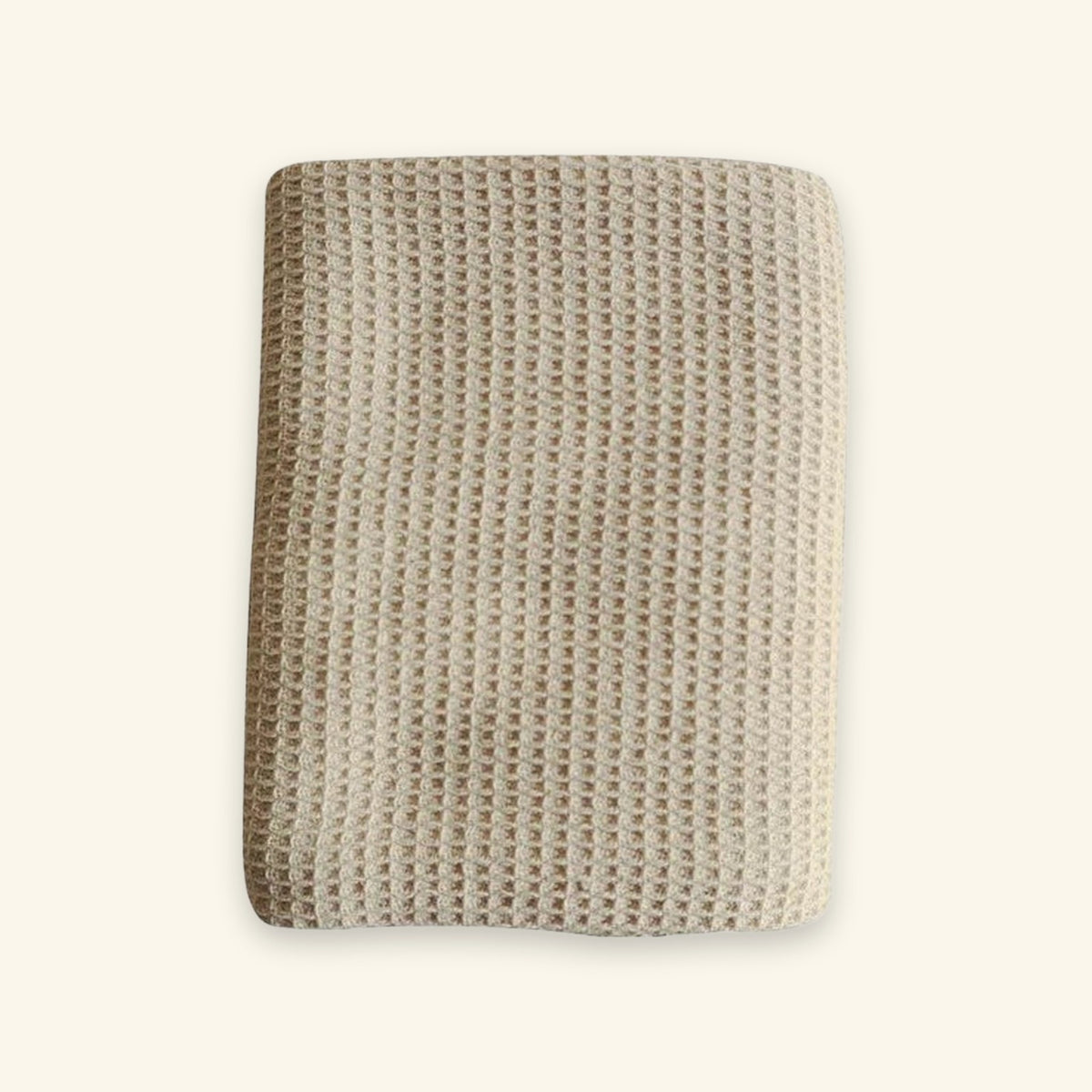 A folded beige textured blanket with a waffle or honeycomb knit pattern, shown against a solid off-white background.