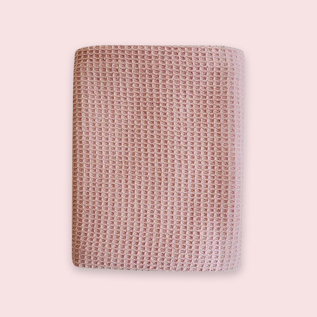 A square pink textured blanket with a waffle or honeycomb pattern, shown flat against a solid pale pink background.