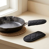 Black quilted oven mitts shown on a beige countertop near a window, with one mitt wrapped around the handle of a dark cast-iron skillet resting on a woven trivet and the other mitt placed beside it.