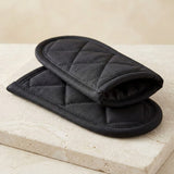 Two black quilted oven mitts stacked on a light beige stone surface against a neutral beige background.