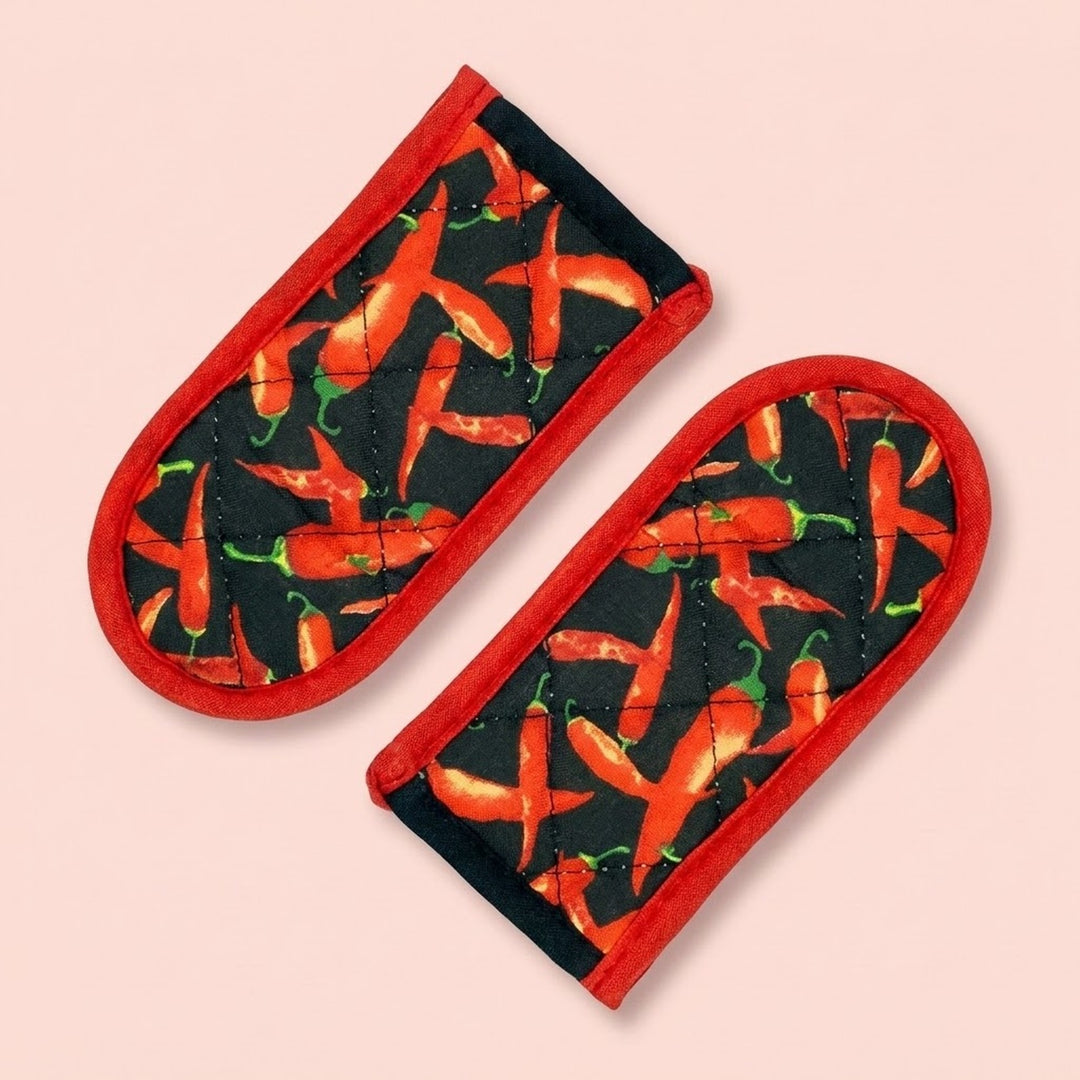 Two quilted fabric oven mitts with a red border and a black background printed with a pattern of red chili peppers, shown on a solid pale pink background.