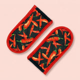 Two quilted fabric oven mitts with a red border and a black background printed with a pattern of red chili peppers, shown on a solid pale pink background.