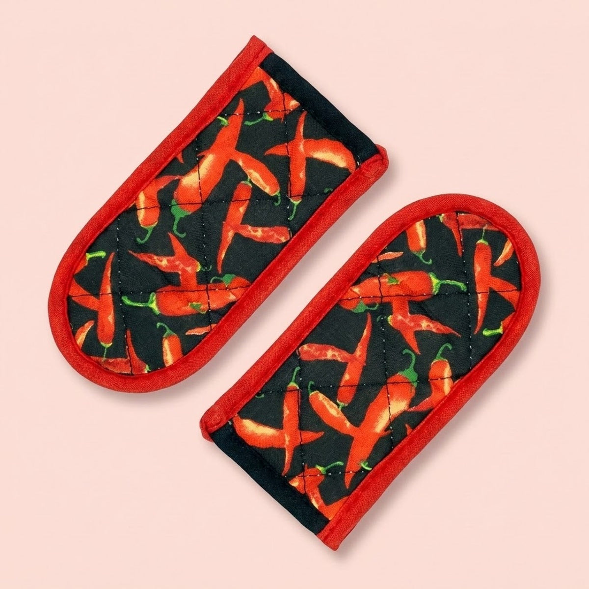 Two quilted fabric oven mitts with a red border and a black background printed with a pattern of red chili peppers, shown on a solid pale pink background.