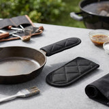 Black quilted sleeves beside a skillet on a gray outdoor table, with one covering its handle and barbecue tools nearby.