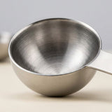 A close-up of a silver stainless steel measuring spoon shows its brushed interior and rounded bowl on a cream surface.