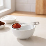 A silver stainless steel measuring spoon marked 5 ml and 1 tsp holds red spice across a white bowl on a sunlit counter.