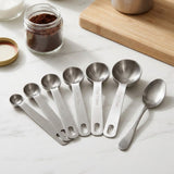 Six silver metal measuring spoons with engraved capacity labels fan across a white countertop beside a teaspoon, coffee jar and wooden board.