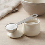 A silver stainless steel measuring spoon filled with white granules rests across a cream ceramic jar and lid on pale wood.