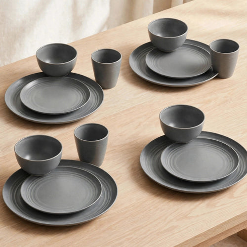 Grey wheat straw dinnerware set arranged as four place settings with plates, bowls, and cups on a light wooden table.