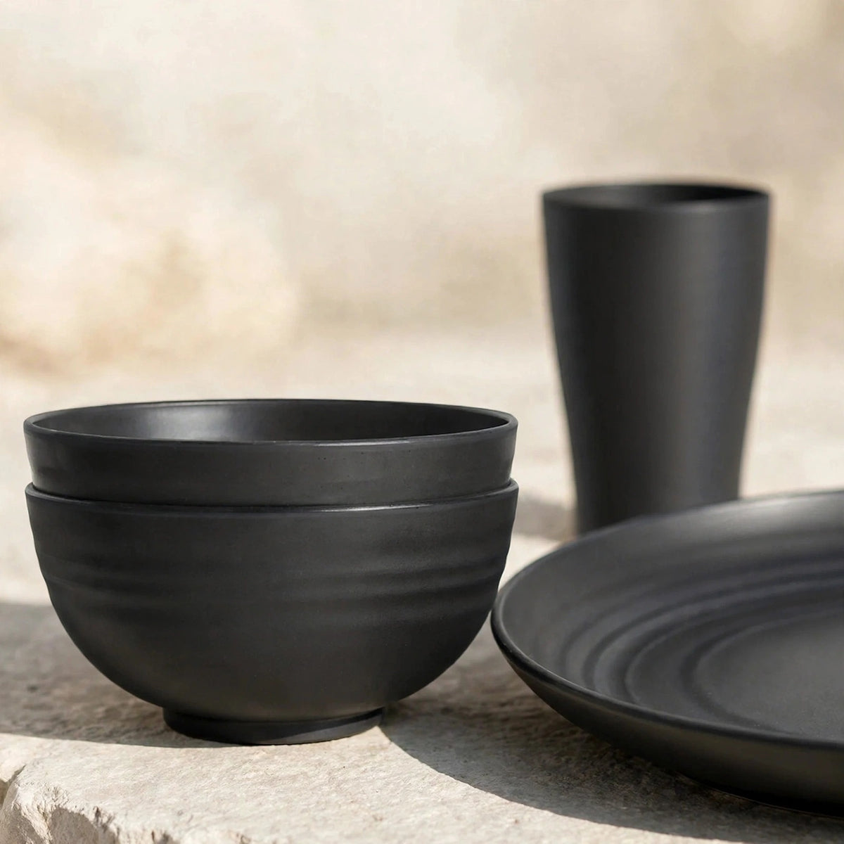 Dark grey wheat straw dinnerware set with two stacked bowls, a plate, and a tall cup on a light stone surface.