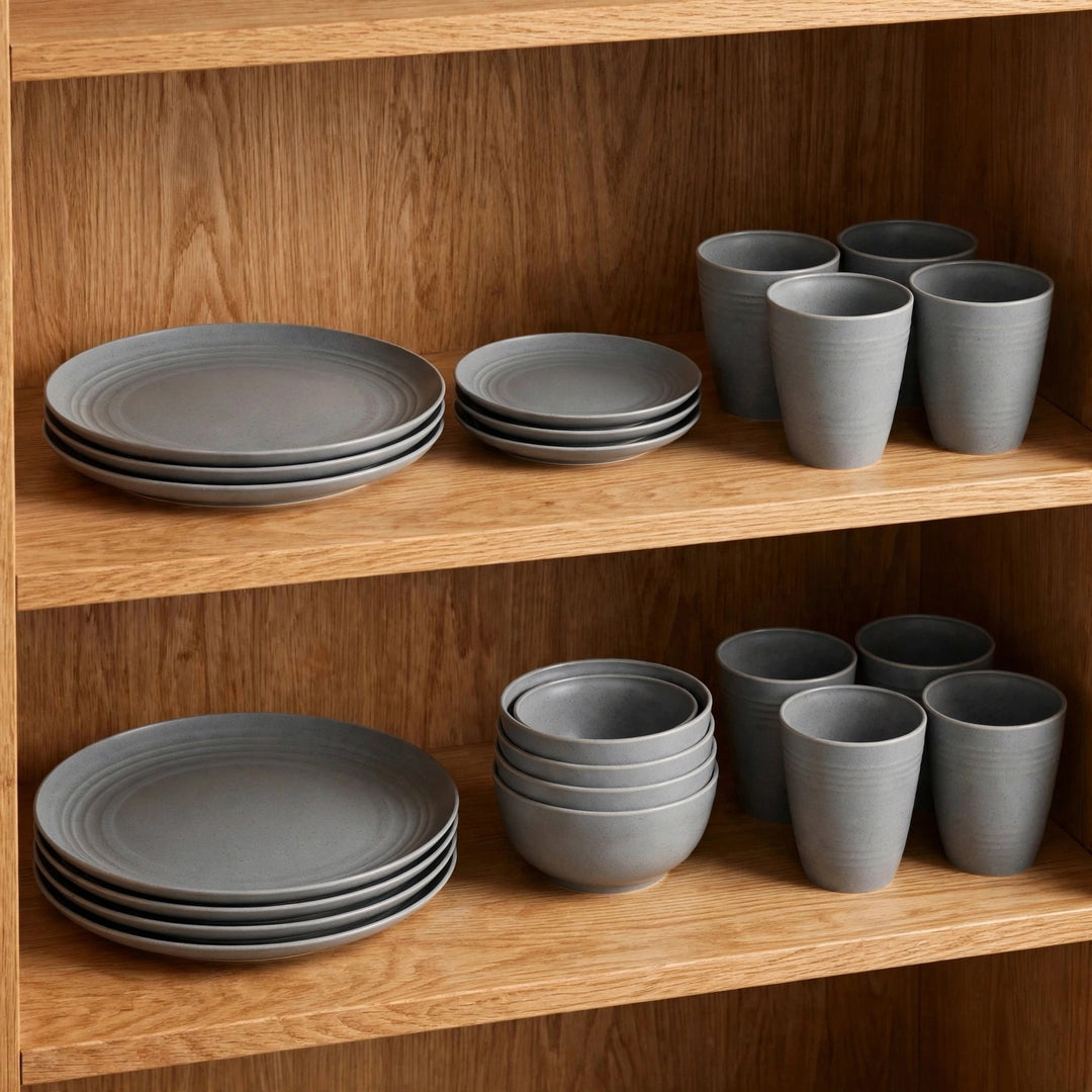 Grey wheat straw dinnerware set with stacked plates and bowls beside matching tall cups on two wooden cupboard shelves.
