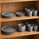 Grey wheat straw dinnerware set with stacked plates and bowls beside matching tall cups on two wooden cupboard shelves.