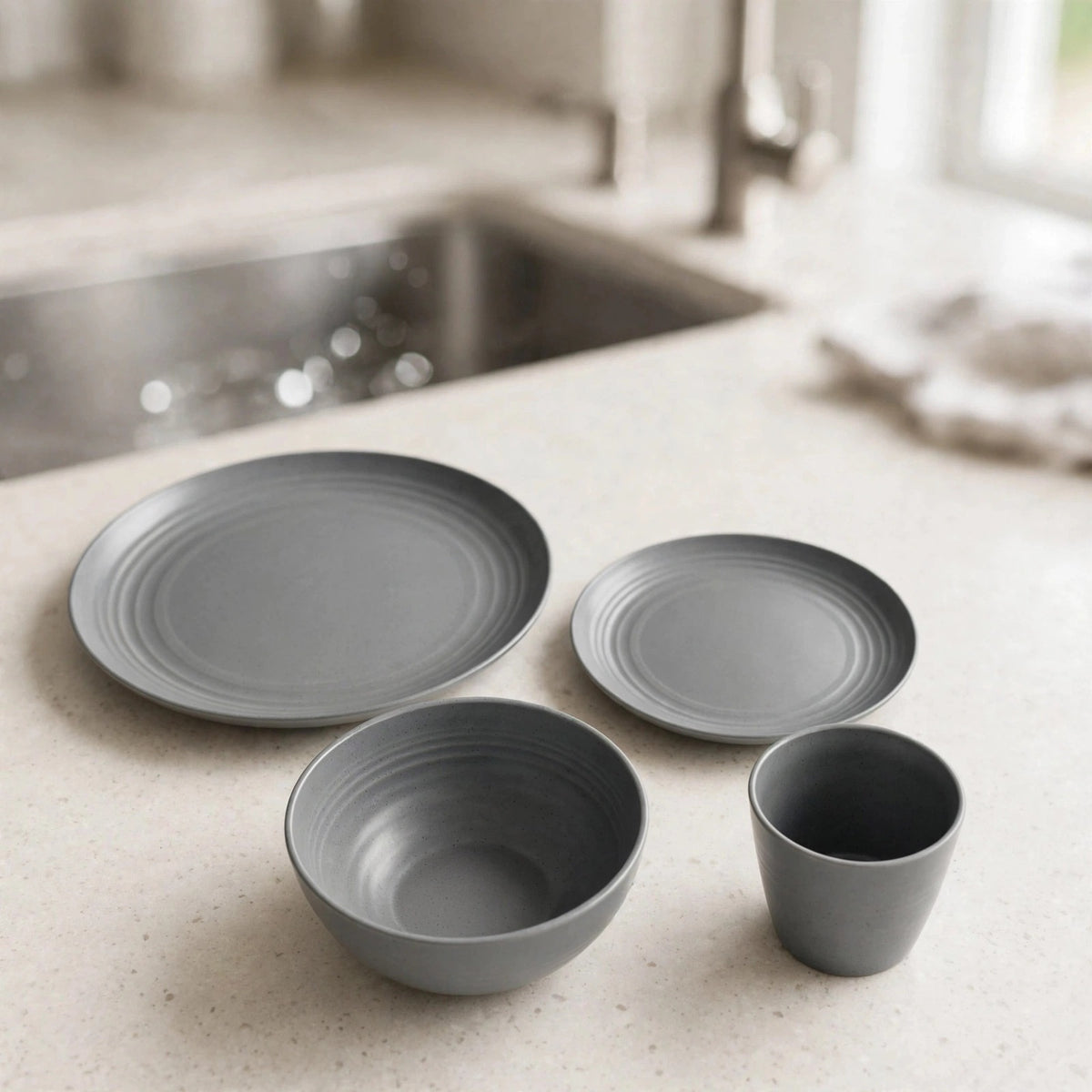 Grey wheat straw dinnerware set with two plate sizes, a bowl, and a cup on a light counter beside a sink.