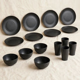 Black dinnerware with ridged detailing includes eight plates, four bowls, and four tall cups arranged on cream fabric.