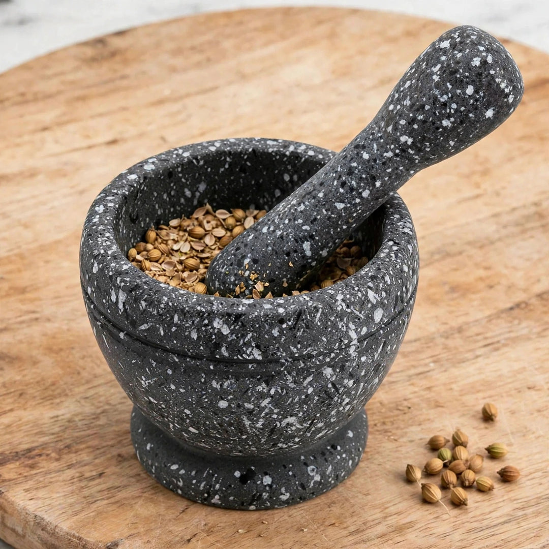 Black mortar and pestle with white speckles holding crushed seeds on a wooden board, with whole seeds scattered beside it.