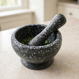 A dark gray speckled stone mortar and pestle set on a light beige countertop, with green herbs visible inside the bowl.