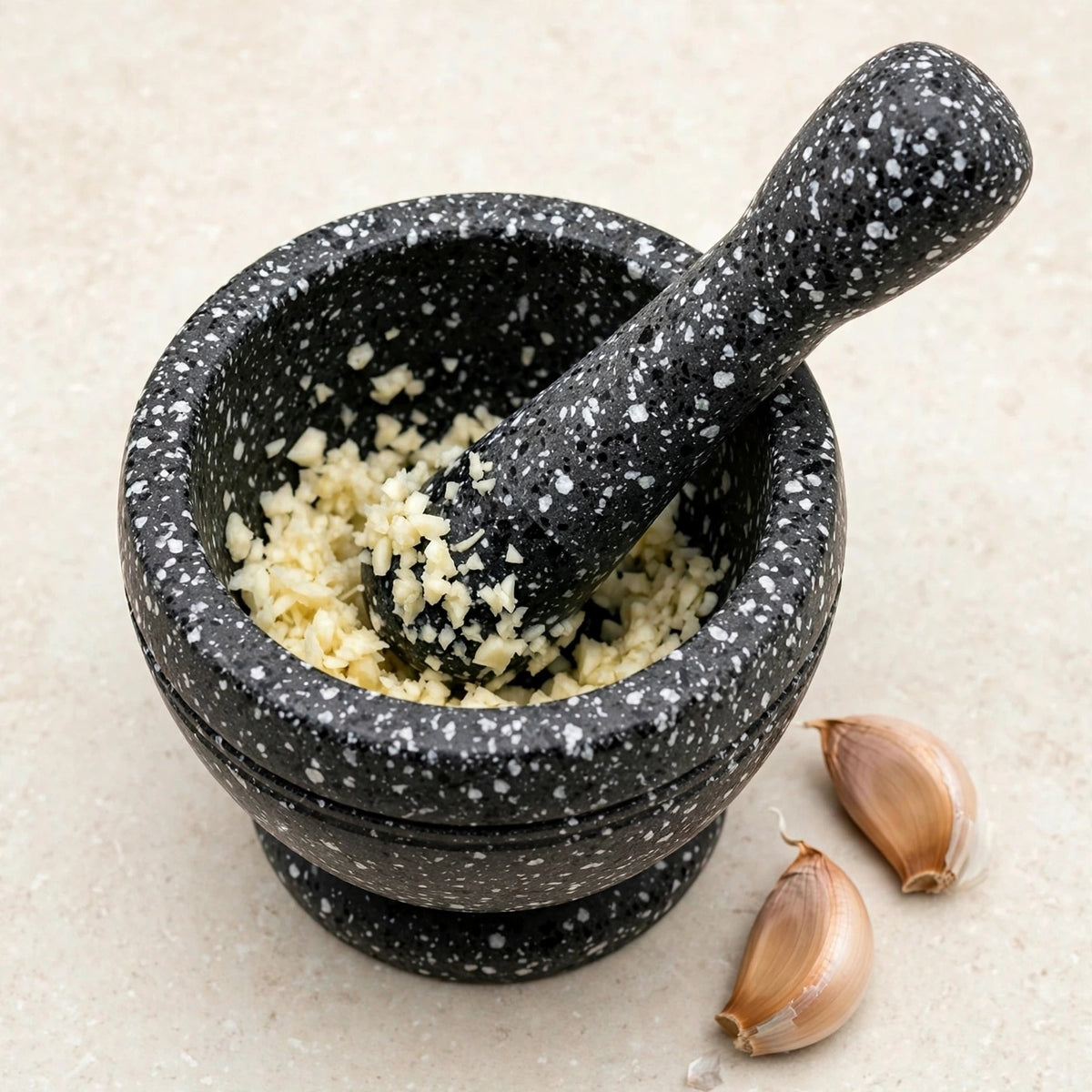 Black mortar and pestle with white speckles containing minced garlic on a pale surface beside two unpeeled garlic cloves.