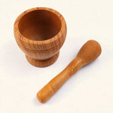 A wooden mortar and pestle set on a plain white background. The mortar is made of medium brown wood with visible grain patterns, and the pestle is placed diagonally next to it.