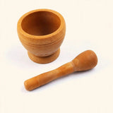A small wooden mortar and pestle set on a plain off-white background. The mortar is a rounded bowl with a short footed base, and the pestle lies diagonally in front.