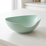 A light mint green speckled ceramic bowl with a unique asymmetrical slant design, resting on a light gray countertop in a bright kitchen setting.