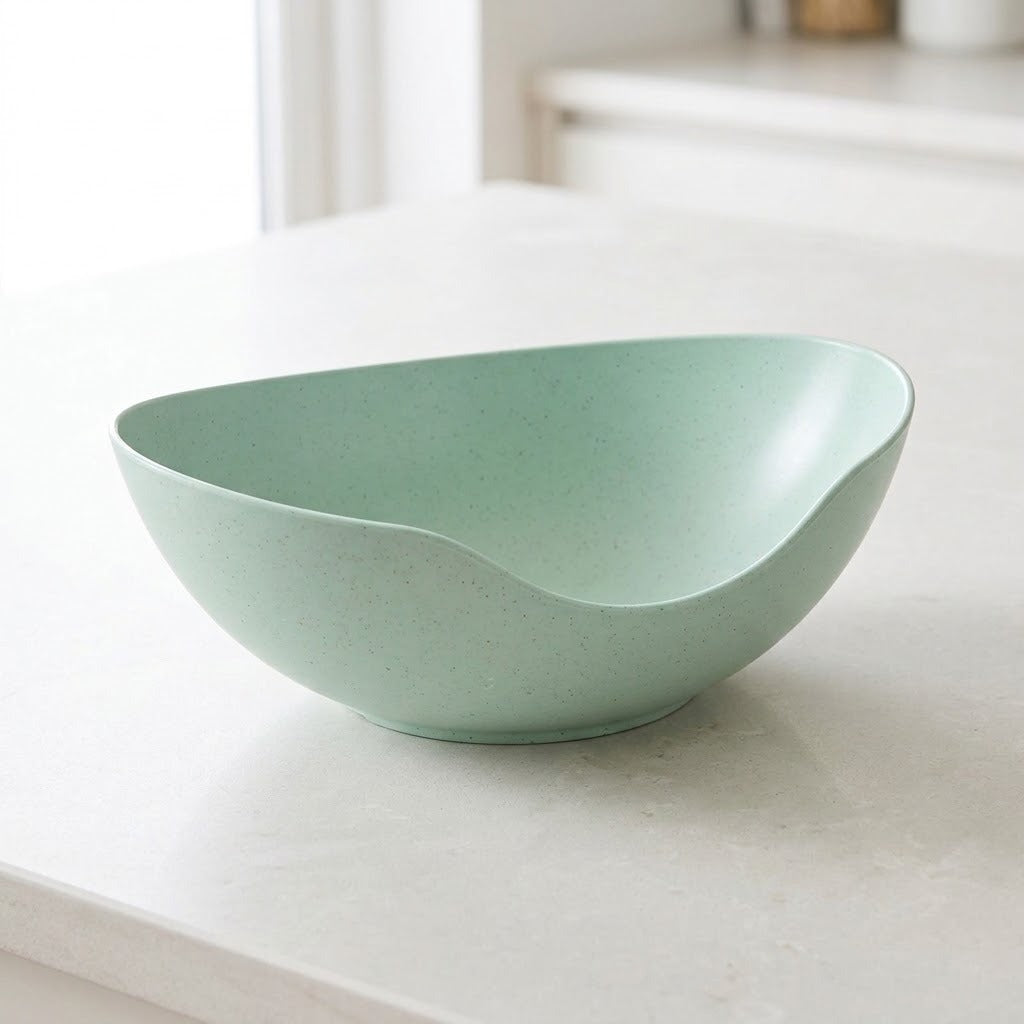 A light mint green speckled ceramic bowl with a unique asymmetrical slant design, resting on a light gray countertop in a bright kitchen setting.