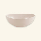 A minimalist, semi-circular ceramic bowl in a light blush pink color with subtle speckled texture, set against a solid cream background.