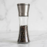 A clear glass pepper mill container filled with whole peppercorns, featuring a brushed stainless steel top, set on a white marble surface with a matching marble background.