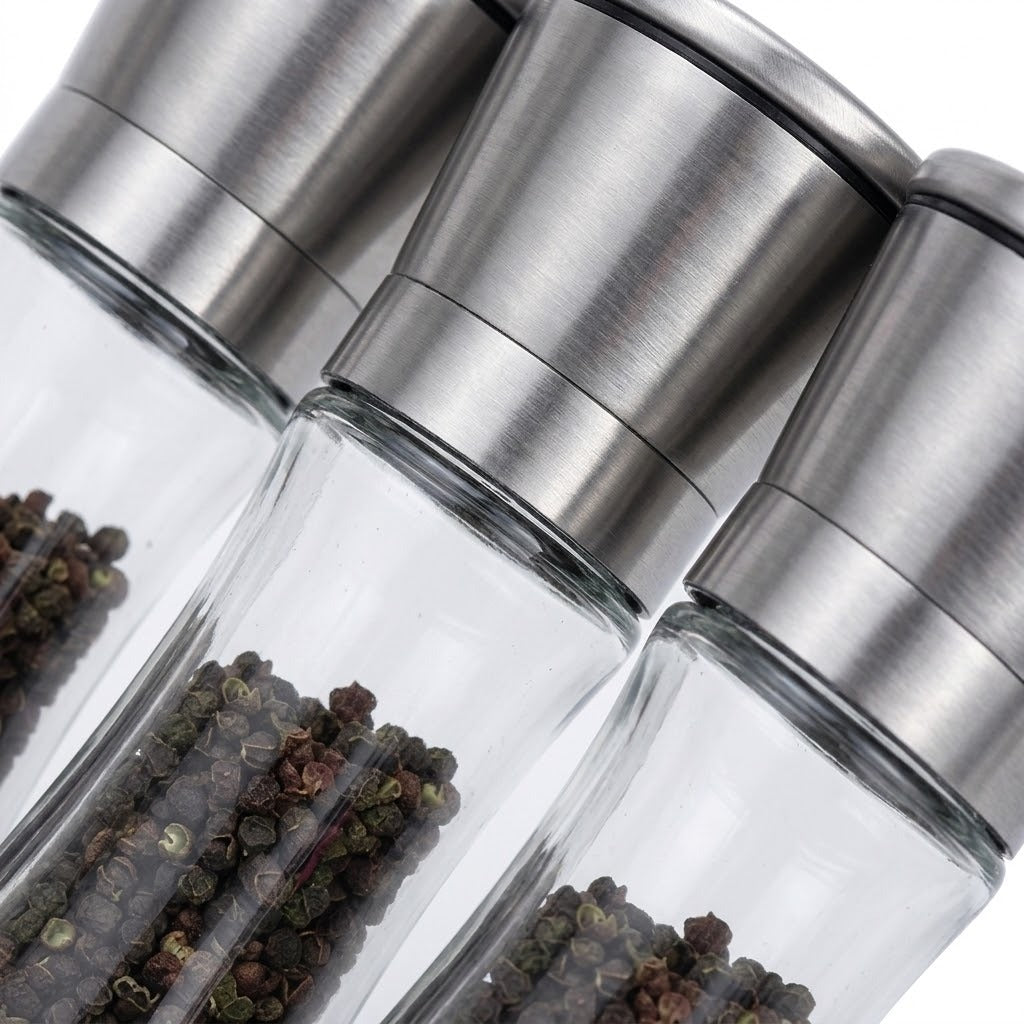 Three clear glass pepper mills with brushed stainless steel caps, shown close-up with peppercorns visible inside, on a white background.