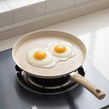 A speckled beige non-stick frying pan with a light wood handle sits on a stovetop burner, cooking two sunny-side-up eggs in a bright kitchen setting.