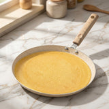 A speckled beige frying pan with a light wood handle sits on a white marble kitchen countertop, filled with a thin yellow crepe or pancake. Sunlight casts soft shadows across the surface, with blurred kitchen items in the background.