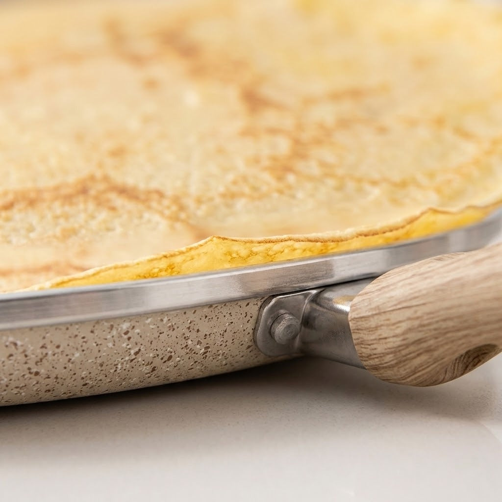 Close-up of a crepe or thin pancake cooking in a speckled beige frying pan with a light wood handle and stainless steel rim.