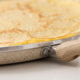 Close-up of a crepe or thin pancake cooking in a speckled beige frying pan with a light wood handle and stainless steel rim.