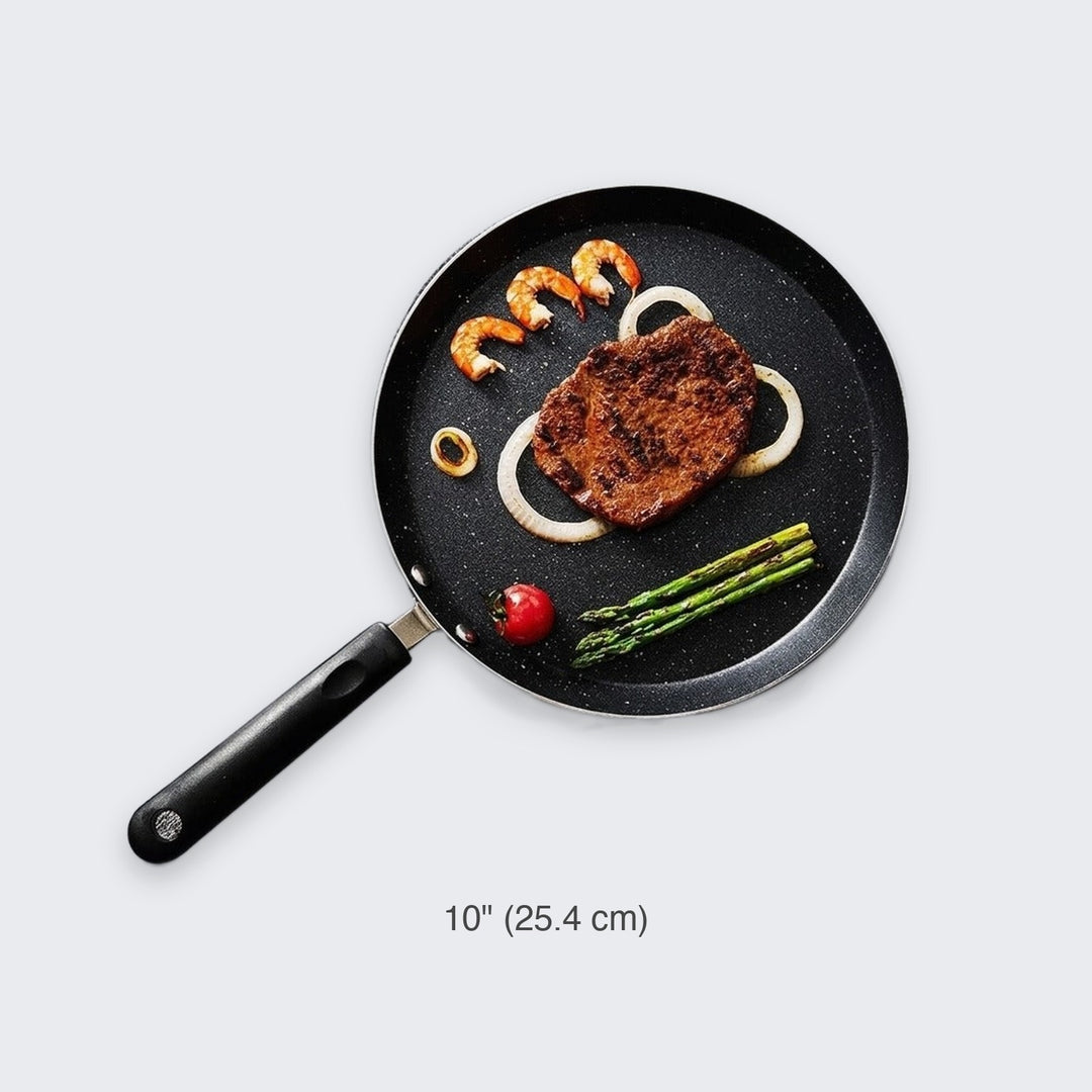A 10-inch black non-stick frying pan with a black handle, shown from a top-down view on a light gray background. The pan is filled with food, including a steak, shrimp, onions, asparagus, and a cherry tomato.