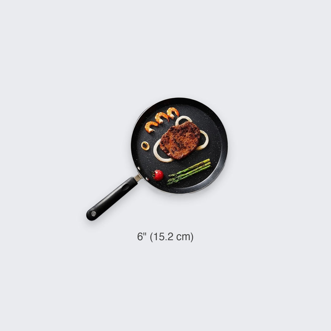 A 6-inch black cast iron skillet with a handle, shown on a light gray background with food items (steak, shrimp, asparagus, onions) cooking inside it and a size label '6" (15.2 cm)' below.