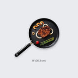 An 8-inch black non-stick frying pan with a black handle, shown from a top-down view on a light gray background. The pan is styled with a steak, shrimp, onion, asparagus, and a cherry tomato.