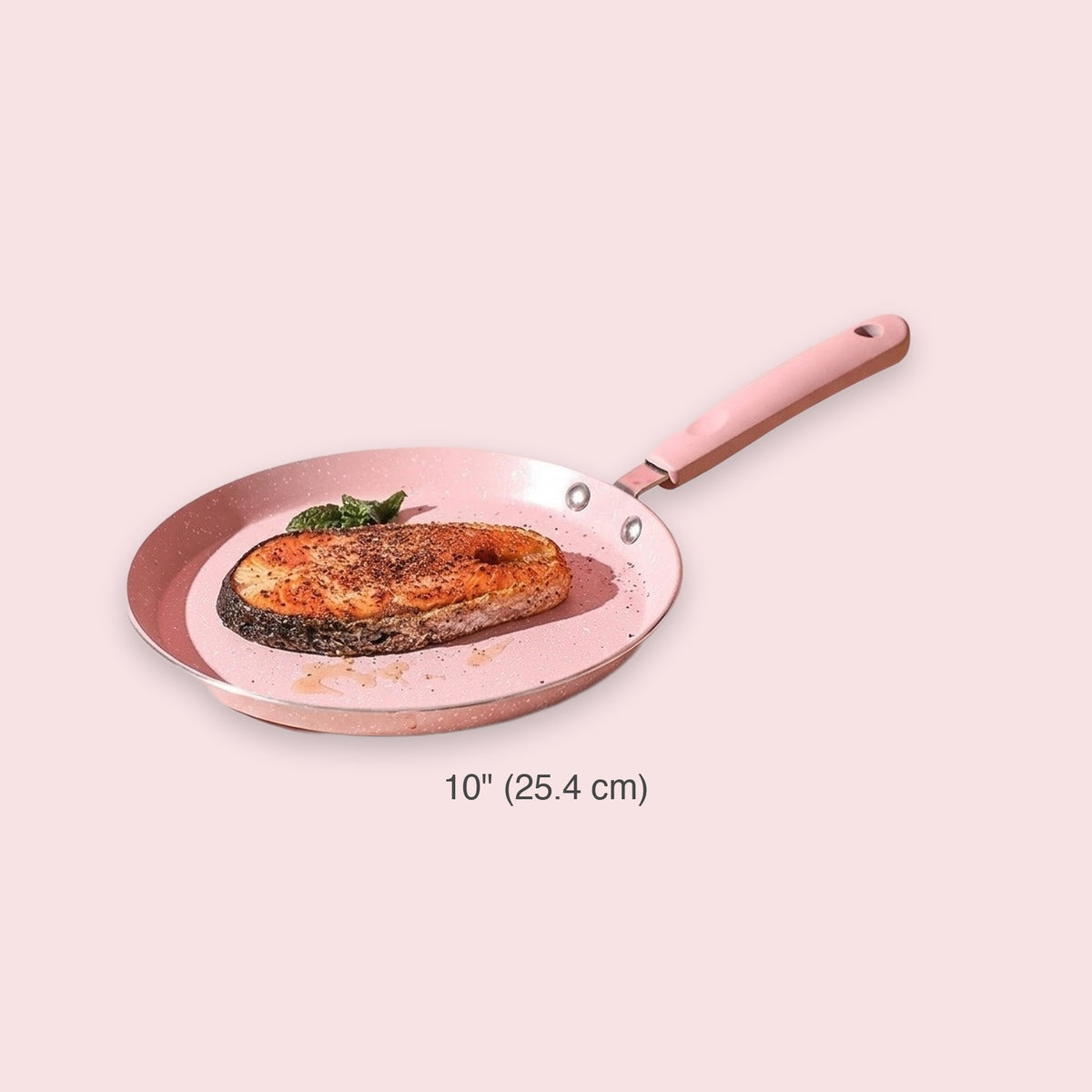 A pink non-stick frying pan with a wooden handle, shown on a pale pink background with a salmon fillet cooking inside. The text '10" (25.