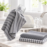 Gray kitchen towels with white stripes and a gray-and-white gingham pattern, displayed on a marble countertop near a sink with stacked plates and glasses in a bright kitchen setting.