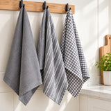 Three kitchen towels hanging from black hooks on a wooden rack: a gray waffle-weave towel, a gray striped towel, and a gray-and-white gingham towel, shown in a bright kitchen setting.