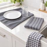 Three gray kitchen towels displayed on a white marble countertop: a waffle-weave placemat with a white plate and clear glass, a folded striped towel, and a plaid towel draped over the sink.