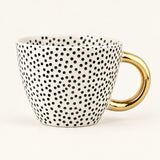 A white ceramic coffee mug covered in small black polka dots, featuring a shiny gold handle, set against a plain off-white background.