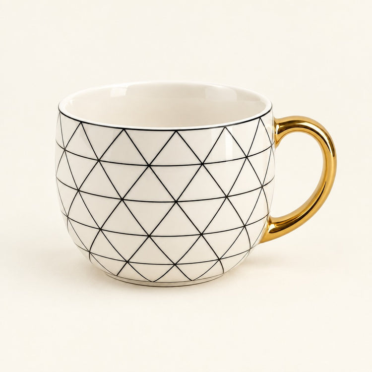 A white ceramic coffee mug with a black geometric diamond pattern and a shiny gold handle, set against a plain cream background.
