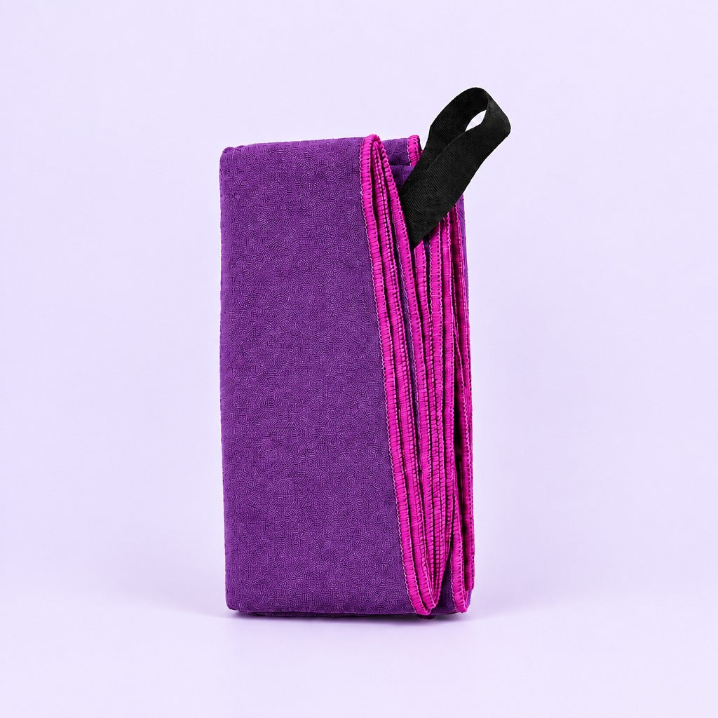 A folded purple yoga block with a black carrying loop, standing upright on a light lavender background.