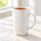 A tall white ceramic mug filled with tea sits on a light marble countertop near a window, with soft natural light casting shadows.