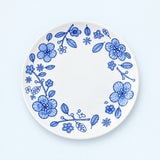A round white ceramic plate with a delicate blue floral border pattern, photographed from directly above on a light blue background.